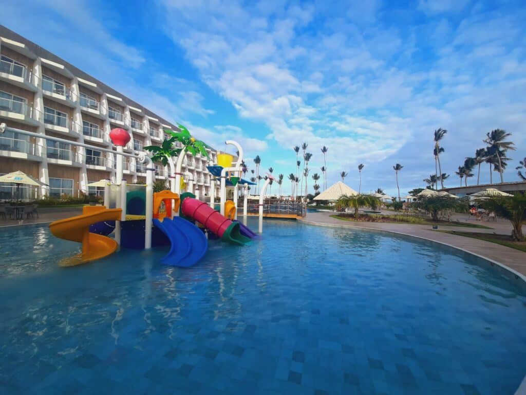 Piscina do Maragogi Brisa Exclusive, com brinquedos para crianças com tobogãs, árvores e à esquerda tem o prédios com sacadas em todos os quartos