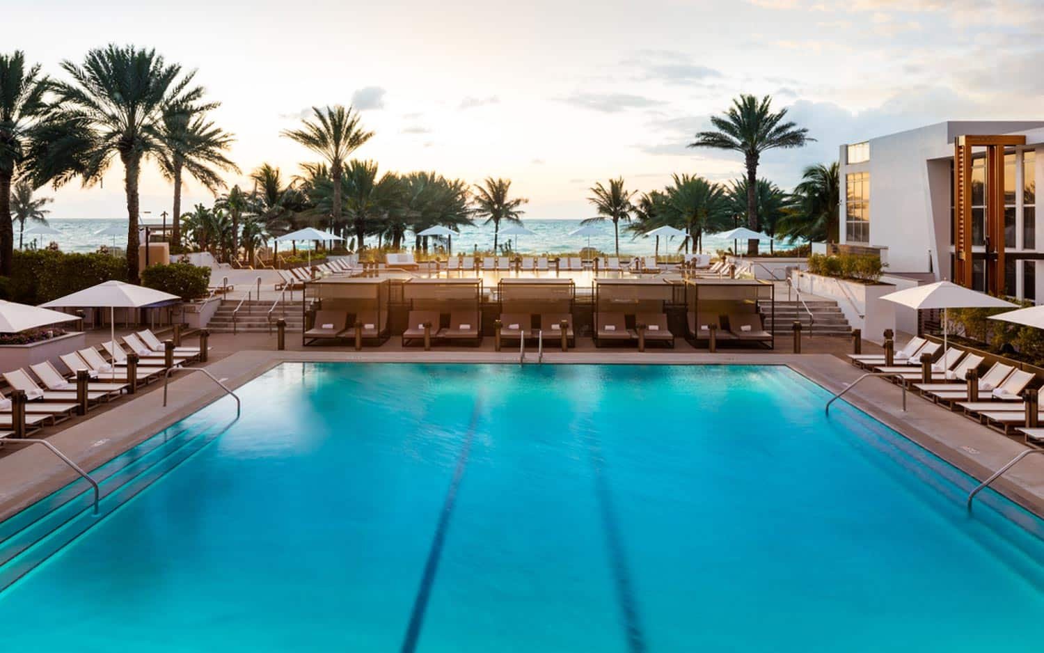 A imagem mostra uma luxuosa área de piscina ao ar livre no Eden Roc Miami, um dos hotéis em Miami Beach indicados. A piscina retangular é cercada por espreguiçadeiras brancas com guarda-sóis. Além da piscina, há uma plataforma elevada com mais espreguiçadeiras e cabanas privadas. Palmeiras altas decoram o ambiente, e ao fundo, é possível ver a praia e o oceano, com o sol se pondo, criando um cenário deslumbrante e tranquilo. A arquitetura moderna do resort complementa o ambiente sofisticado e relaxante.