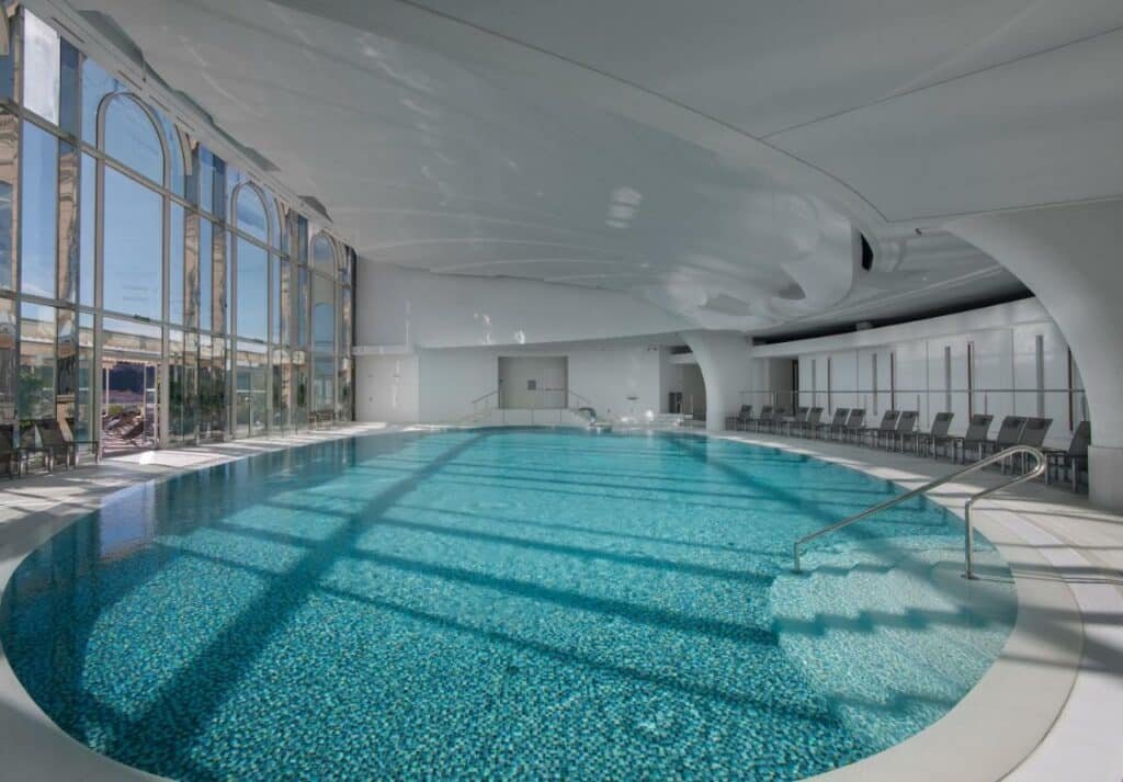 Piscina do Hôtel Hermitage Monte-Carlo. Piscina na área coberta, ao redor cadeiras de tomar sol. No lado direito, uma parede de vidro com vista para a cidade.