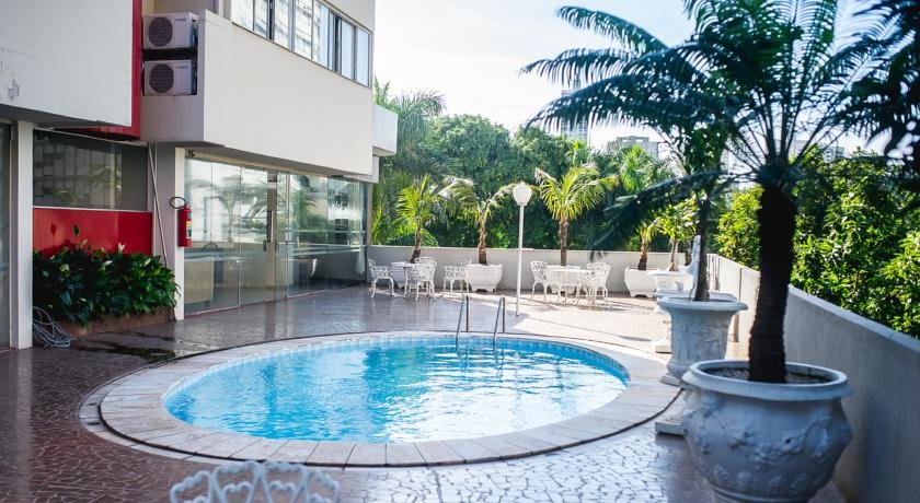 Piscina redonda rodeada por árvores e algumas mesas com cadeiras. No canto é possível ver uma parte do hotel.