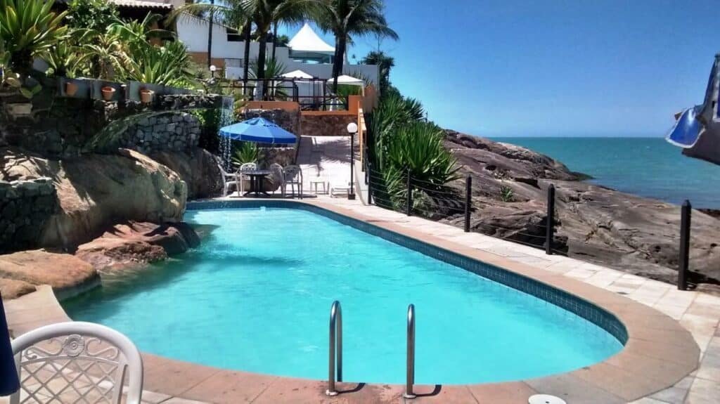 Piscina ao ar livre do Hotel Pontal das Rochas. Pedras no anto esquerdo, uma piscina no meio, e pedras no canto direito e o mar. Atrás uma mesa com guarda-sol, cadeiras e a rampa com acesso para o hotel.