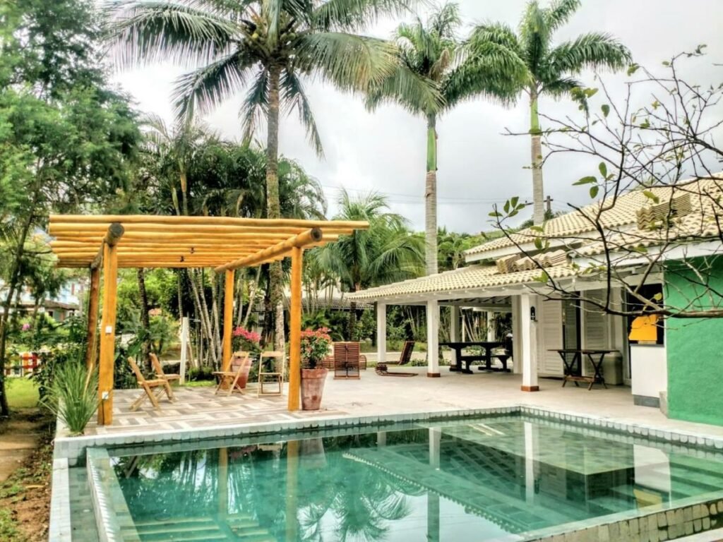 Piscina da Linda Casa Praia de Pauba em condomínio durante o dia com piscina a frente, atrás da piscina uma área de lazer e do lado direito da piscina ao fundo a propriedade. Representa airbnb na praia de Calhetas.