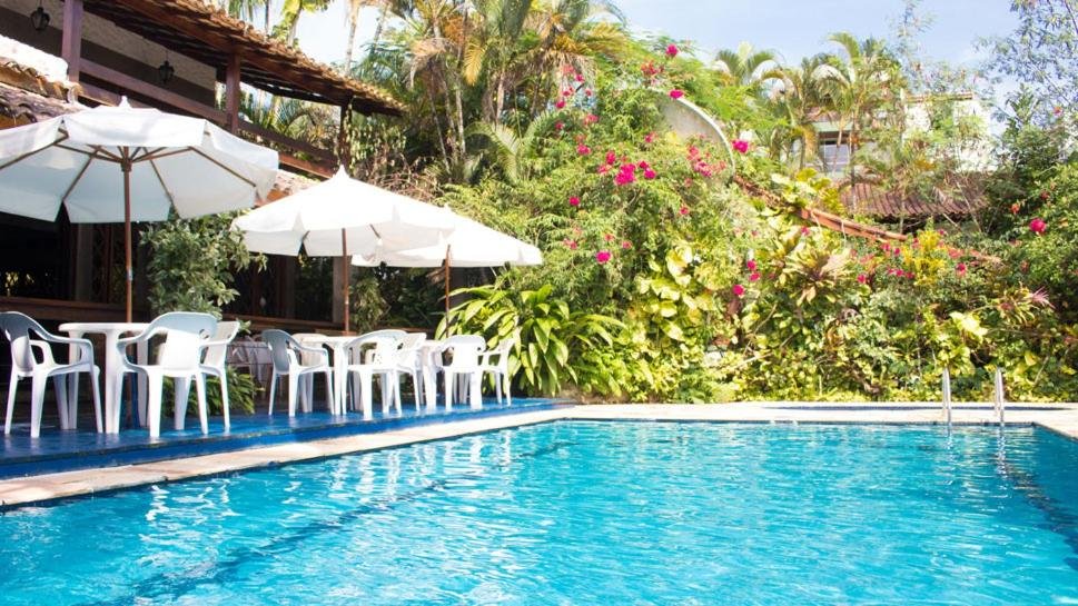 Piscina da Pousada Albatroz, bem ampla, cercada por vegetação com flores e árvores, do lado esquerdo, é possível ver cadeiras e guarda-sóis