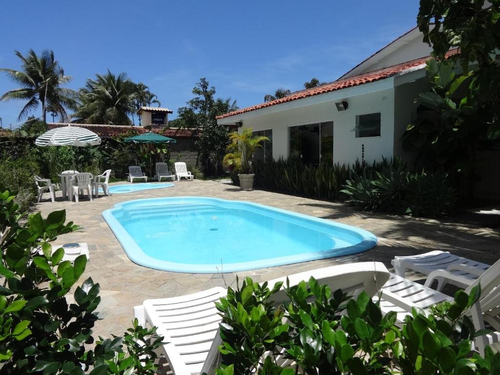 Piscina da Pousada Baía das Conchas,, cercado por plantas e arbustos, algumas cadeiras, mesas e guarda-sóis ao redor