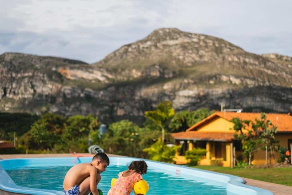 área da piscina com duas crianças morenas brincando na beirada, uma menino e uma menina. Ao fundo, no canto direito, é possível enxergar a pousada amarela e, mais atrás, as montanhas e pedras da região.