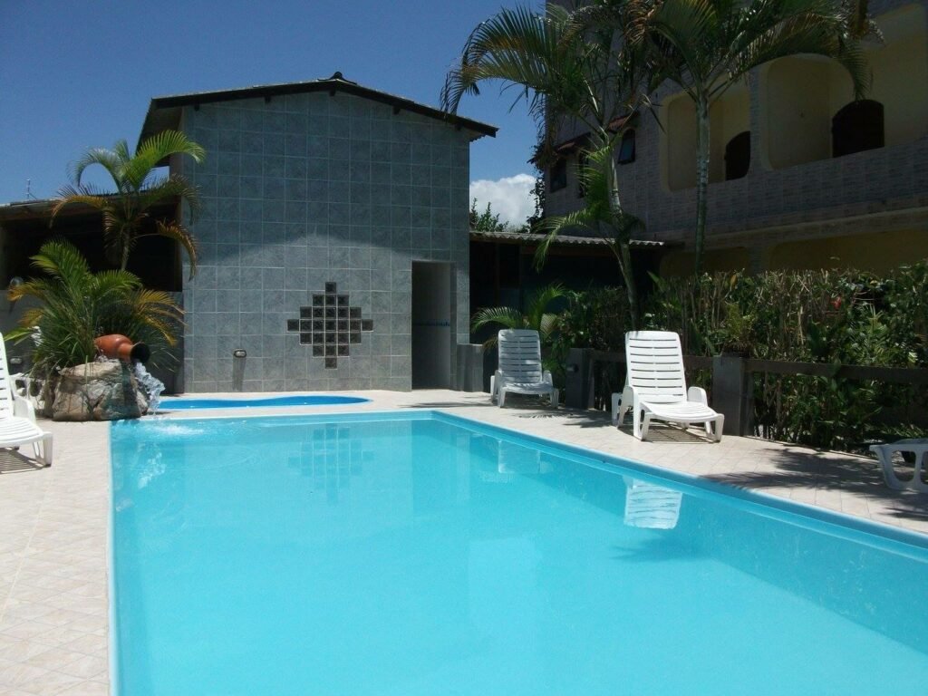 Piscina ao ar livre, da Pousada Boramar. Uma piscina grande e outra piscina infantil pequena. Ao redor cadeiras de tomar sol e atrás o espaço do banheiro. Do lado direito a pousada.