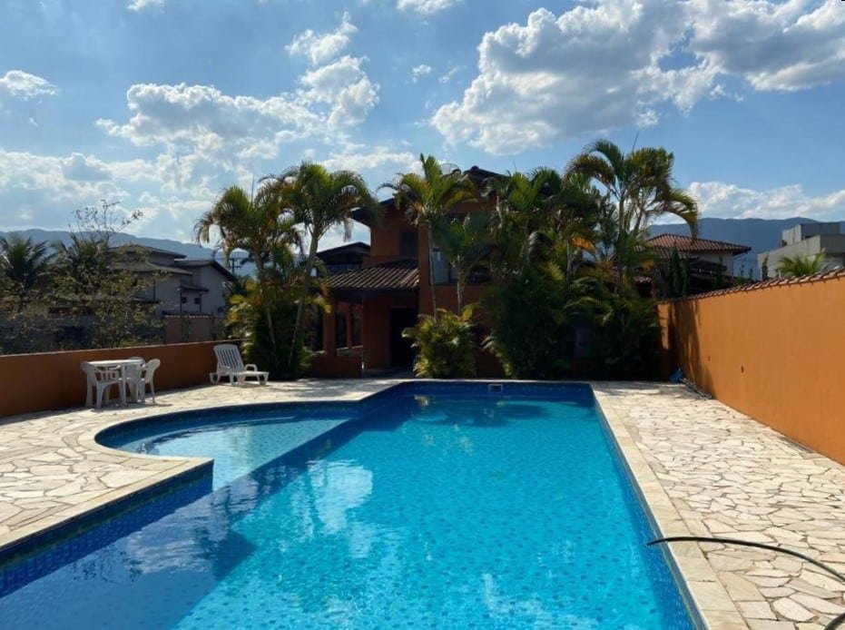 Uma piscina grande ao ar livre na Pousada Familiar Morada do Rapha. No fundo, a pousada, com várias árvores e bastante sombra.