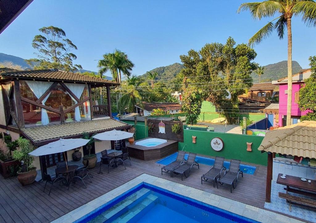 Vista de cima da piscina da Pura Vida Maresias com cadeiras perto da borda da piscina, no lado direito, área de churrasqueira, lado esquerdo, banheira de hidromassagem, cadeiras com mesa e guarda-sóis e a hospedagem atrás.