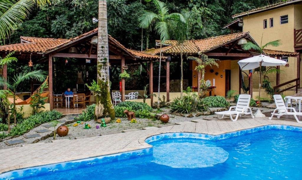 Vista da piscina da Pousada Recanto dos Tangaras durante o dia com cadeiras brancas do lado direito e ao fundo estrutura com cobertura com mesas e churrasqueira. Representa pousadas na praia do Engenho.