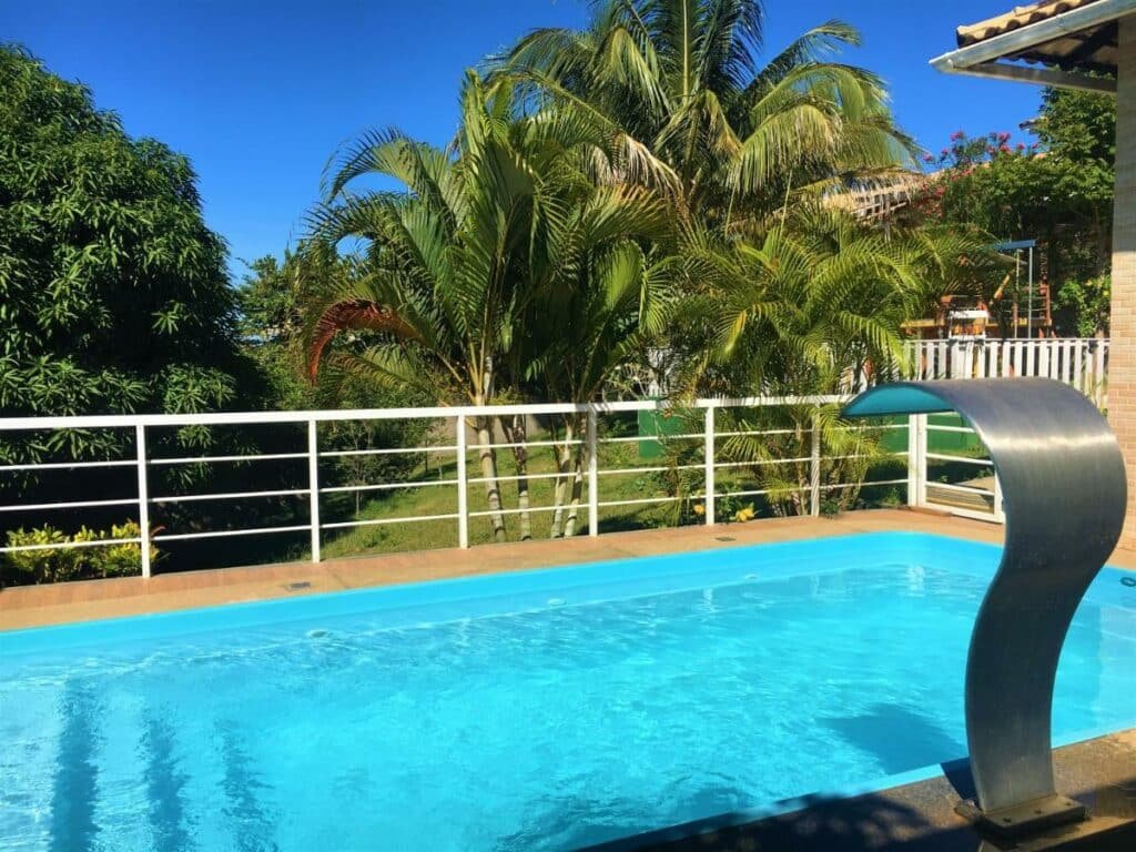 Piscina ao ar livre da Pousada Santa Helena. Uma cascata no lado direito, e atrás da piscina uma grade de apoio e atrás árvores.