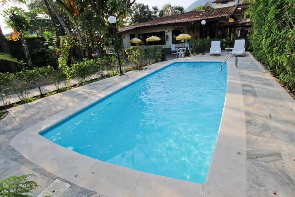 Piscina na Pousada Vivenda do Flamboyant, bem ampla, com muitos arbustos dos dois lados, guarda-sóis, cadeiras e alguns postes de luz