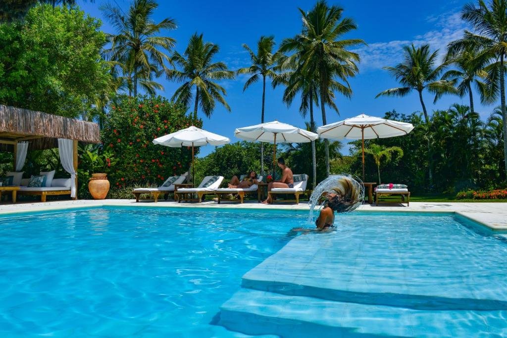 Vista da piscina do Hotel e Resort Villas de Trancoso durante o dia com uma mulher loira dentro da piscina. Ao fundo tem um casal conversando sentado nas cadeiras próximo a piscina. Representa Resorts em Trancoso.