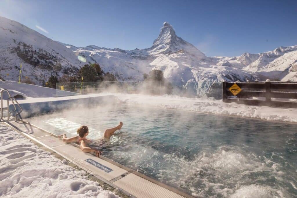 Piscina aquecida ao ar livre do Riffelalp Resort 2222m durante o dia com piscina quadrada no centro com uma mulher dentro da piscina e que oferece vista para as montanhas.