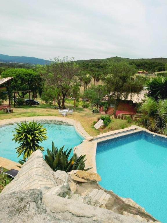 piscinas da Sinhá Cipó com duas áreas de banho separadas por uma pedra. Há várias árvores e construções ao redo da piscina e ao fundo muitas montanhas verdejantes.
