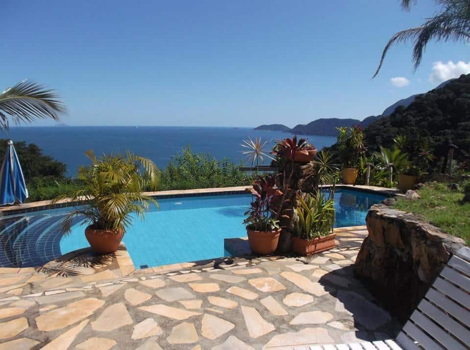 Vista da piscina do Vila Lia durante o dia com vasos de plantas em volta e ao fundo piscina. Representa pousadas na praia de Guaecá.