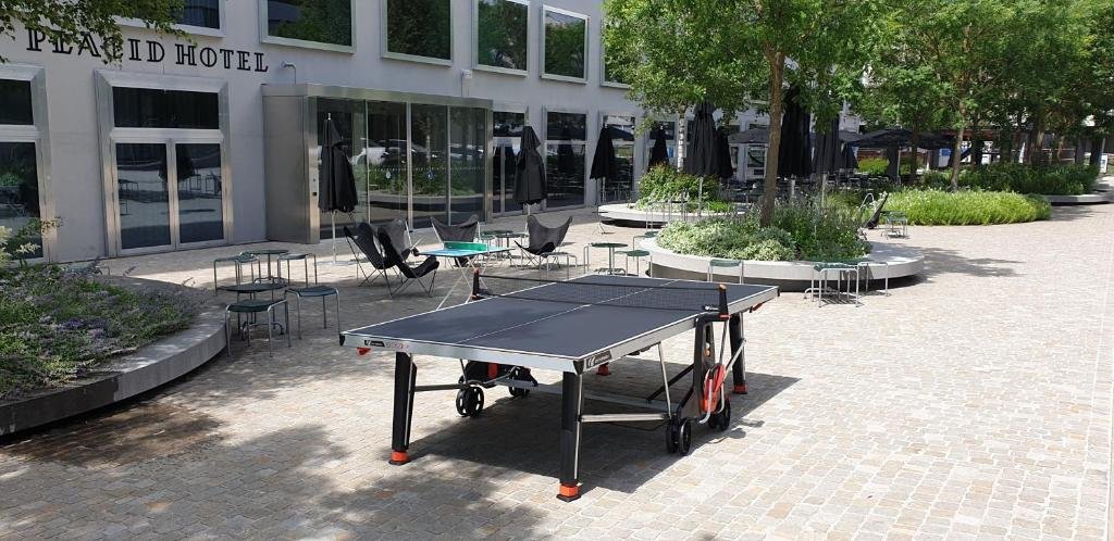 Área externa do Placid Hotel Design, com portas de vidro dando acesso ao interior do local, mesa de ping-pong, mesa com cadeiras e jardins com árvores e folhas verdes