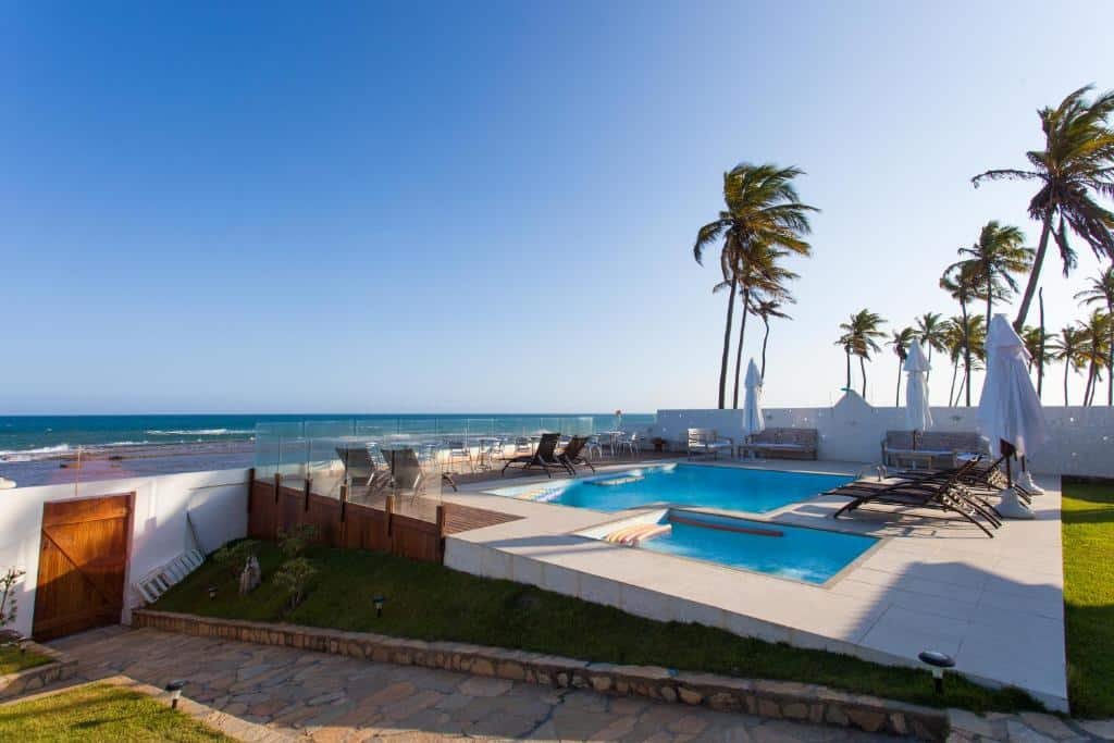 Piscina da Pousada a Capela. Uma piscina no lado direito, com cadeiras de tomar sol ao redor. Na frete um corredor com o portão da pousada, no lado esquerdo a praia.