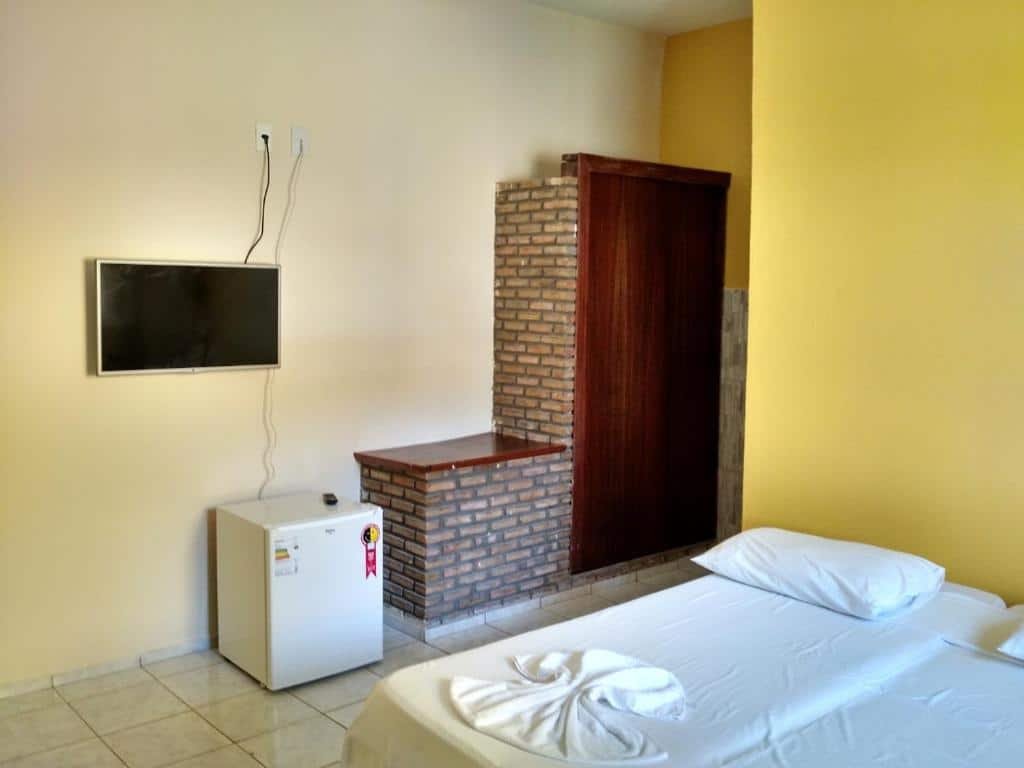 Quarto da Pousada Águas do Piruí. Uma cama de casal no lado direito, no lado esquerdo o guarda-roupa, um banco de apoio de tijolinhos, uma televisão e um frigobar.
