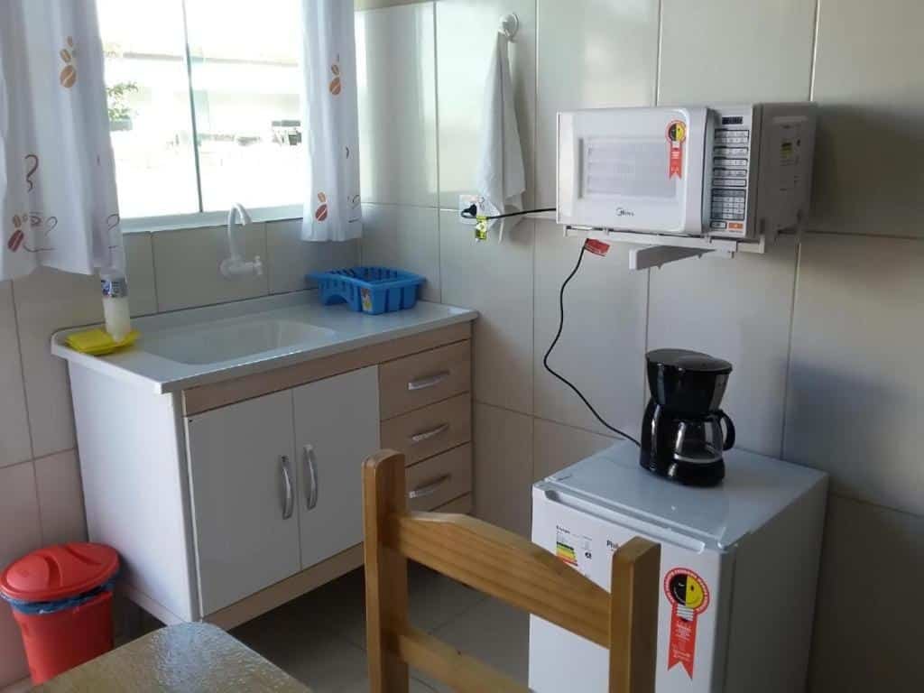 Cozinha da Pousada Ancoradouro. Uma pia está encostada na parede e abaixo de uma janela. Ao seu lado está uma pequena geladeira, uma cafeteira e um microondas. No canto da imagem é possíver também ver uma mesa com cadeira.