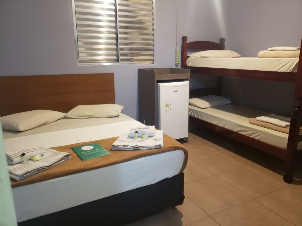 Quarto quádruplo da pousada Beira Mar. Há uma cama de casal, um frigobar e uma beliche com duas camas de solteiro. Há toalhas e produtos de higiente em cima da cama de casal.