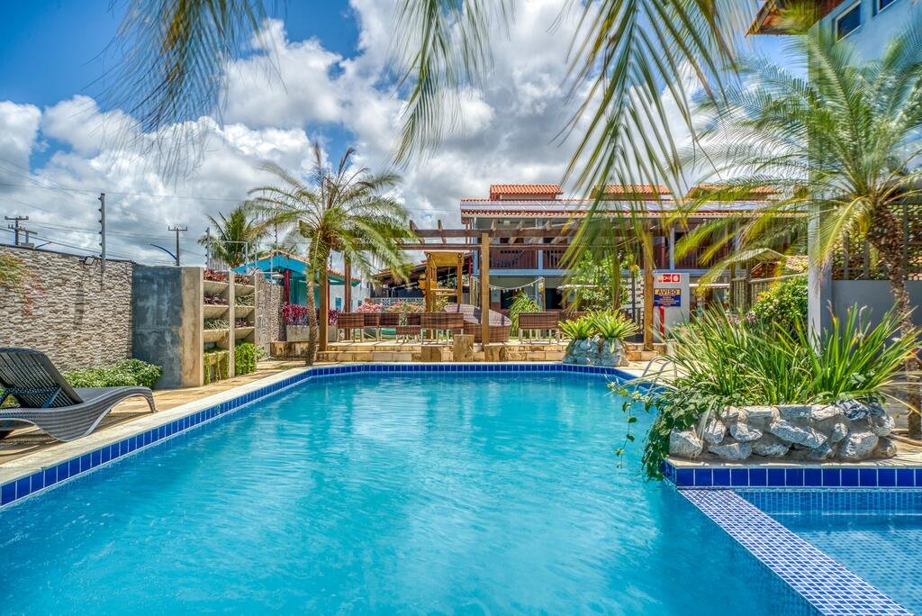 Piscina da Pousada Blue Viking, com algumas pedras com folhas verdes decorando o ambiente, além de algumas árovres, espreguiçadeiras e cadeiras em volta para ilustrar as pousadas em Alagoas