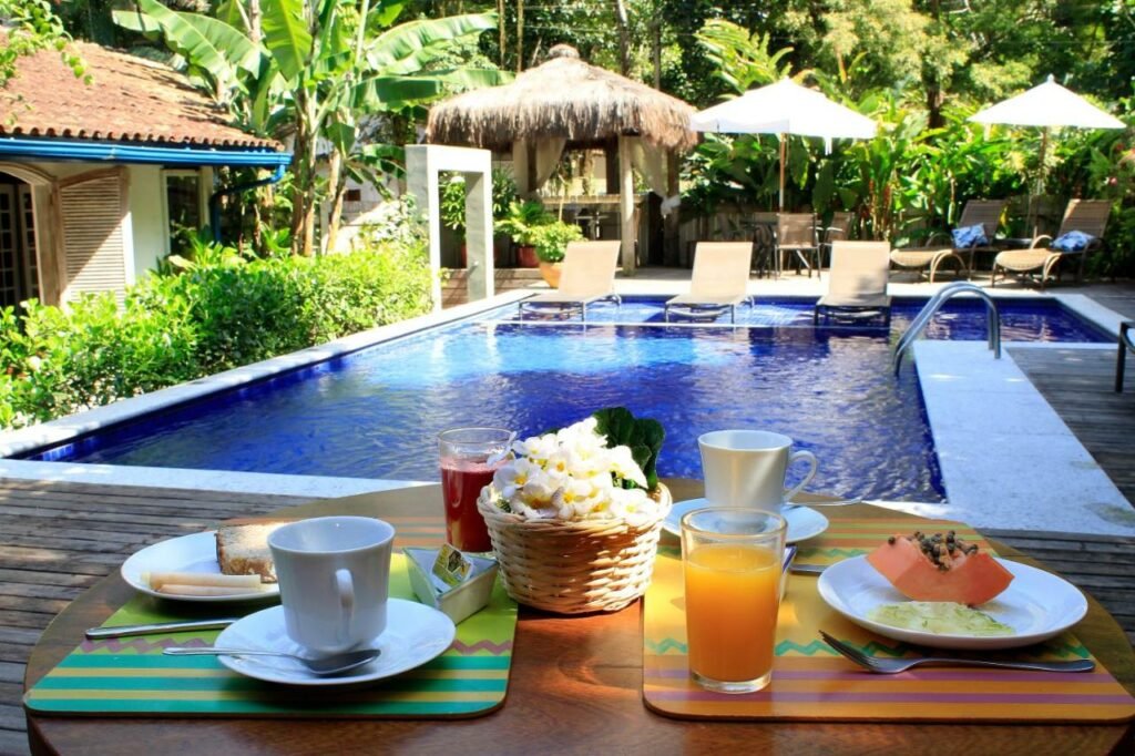 Área externa da Pousada Canto do Camburi. Uma grande piscina com espreguiçadeiras na área rasa ocupa a maior parte da imagem. Em uma mesa perto da piscina estão dois pratos com café da manha, sucos e xícaras. Ao redor estão vários árvores, guarda-sóis e uma cabana.
