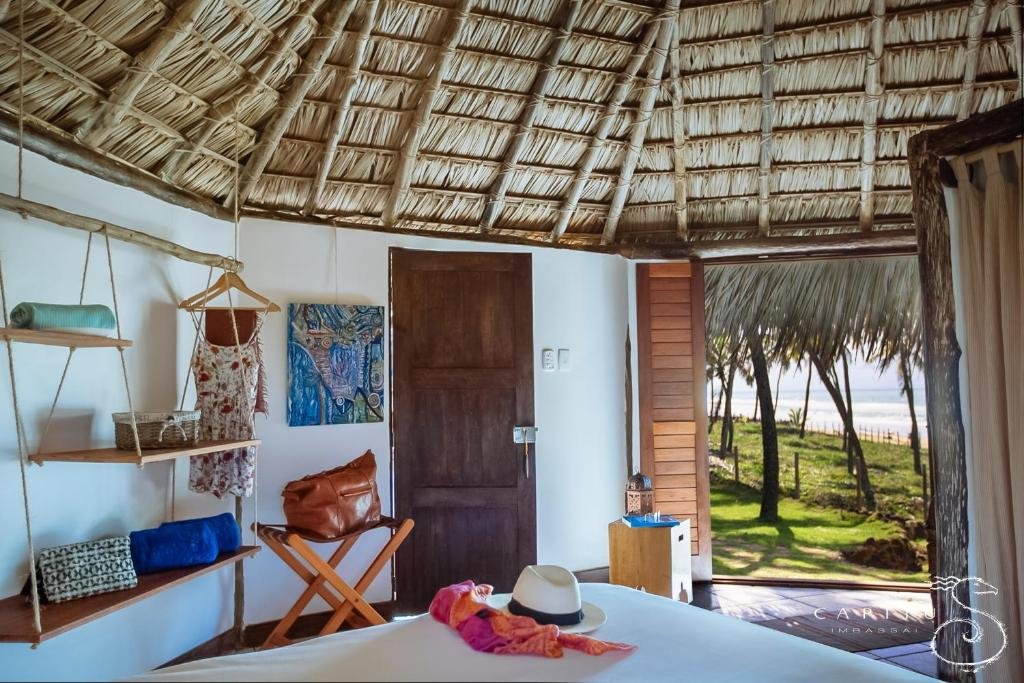 Quarto da Pousada Capitù. Uma cama de casal na frente, no lado direito prateleiras penduradas com corda, com toalhas e mantas, um banco de madeira com uma bolsa, a porta do banheiro e do lado esquerdo a porta do quarto com vista para a praia.