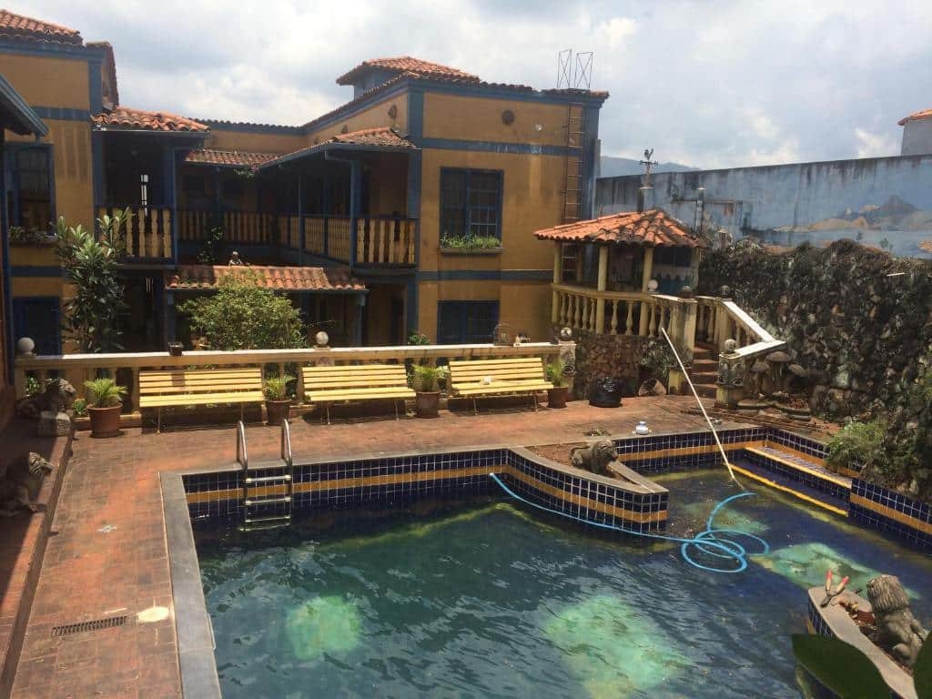 área da piscina da Pousada Casarão da Pedra. Há três bancos de praça amarelos dispostos lado a lado em frente a piscina. Mais atrás é possível enxergar os dois andares da construção antiga, pintada em amarelo e azul
