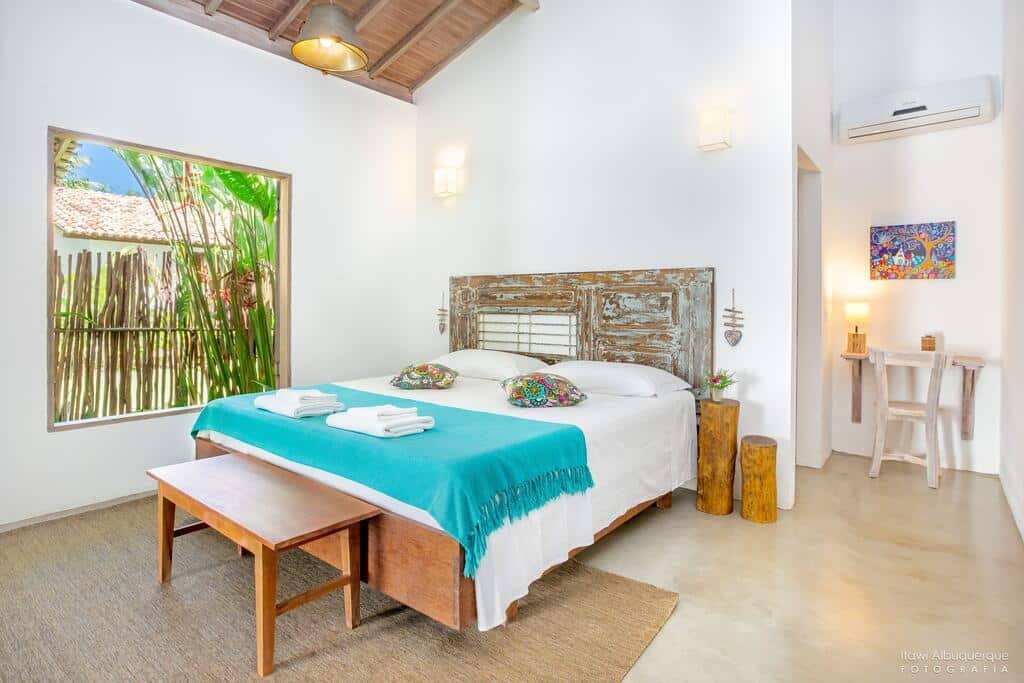 Quarto da Pousada Casotas com cama de casal e janela com vista de uma parede de bambus e folhas verdes