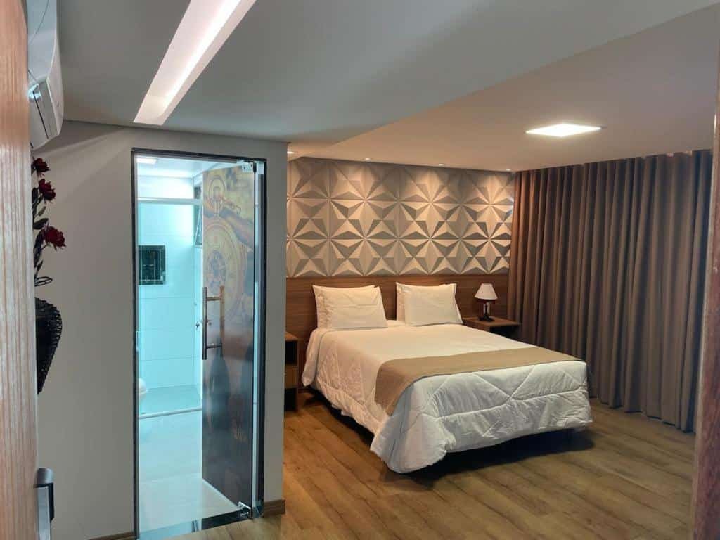 quarto da Pousada Circuito dos Inconfidentes em Congonhas com uma parede decorada com desenhos geométricos servindo de cabeceira para a cama de casal. No lado esquerdo da cama há uma parede coberta por pesadas cortinas e no lado direto há uma porta de vidro aberta, com uma adesivo de relógio cobrindo-a inteira, e pode-se enxergar o boxe do banheiro
