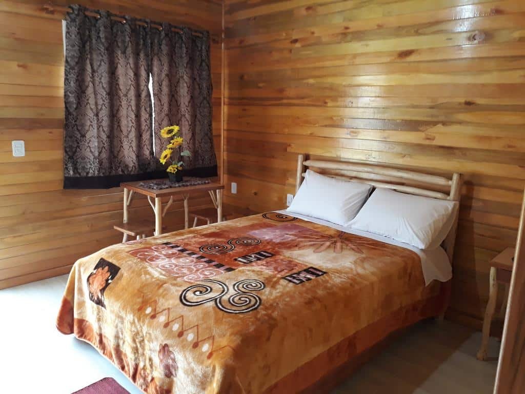 Quarto da Pousada da Gruta, todo em madeira, com janela ao lado da cama com cortinas. Abaixo da janela há uma mesinha com vaso de girassóis ao lado da cama de casal bem ao centro da mesa. Do outro lado do quarto há mais uma mesinha de madeira.