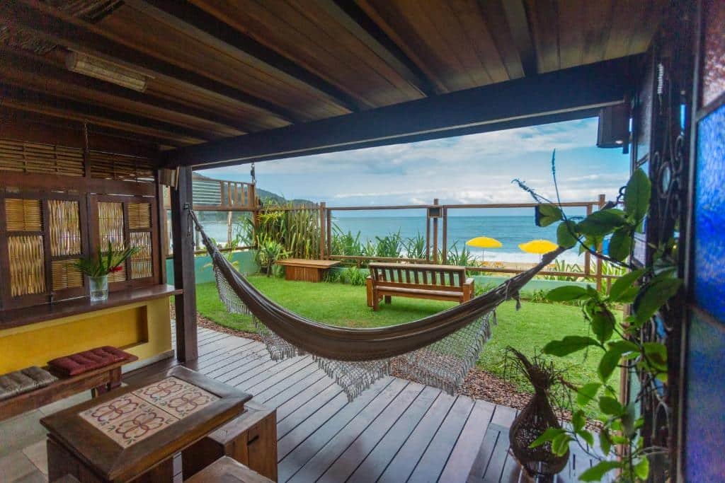 Área coberta e área externa da Pousada DiMari. Na área coberta, que está em primeiro plano, rdtão alguns bancos, uma mesinha e uma rede. Ao fundo está um gramado com um banco de frente para a vista da praia.