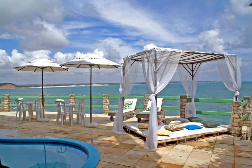 Área da piscina da Pousada Ecobaia. É possível ver a piscina no canto inferior esquerdo e, ao seu lado direito, existem mesas com cadeiras e guarda-sóis. Além disso, existe um espaço para descanso com colchãol amofadas e um dossel. Toda essa região está de frente para o mar. Essa é uma das pousadas em Baía Formosa, RN.