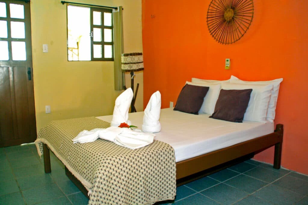 Quarto da Pousada Ecobaia. Uma cama de casal está no meio, ao lado de uma porta e uma pequena janela na mesma parede. Abaixo da janela há um abajur.