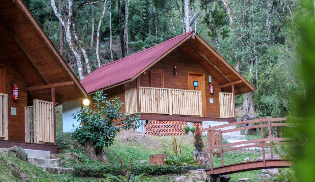 A imagem mostra a fachada da Pousada Encanto do Avencal, um dos chalés na Serra Catarinense, com varanda com vista para a natureza. Tudo é em madeira, desde a estrutura, até porta, janela e resguardo. Há uma ponte madeira indicando um rio mais à frente.
