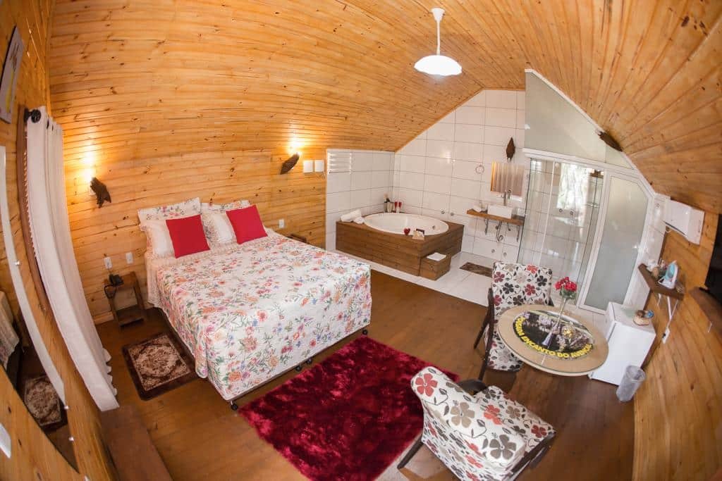 Quarto da Pousada Encanto do Avencal com cama de casal, tapete felpudo em frente além de mesa redonda com duas poltronas. De ambos os lados da cama há uma mesinha com luminárias. Do outro lado, é possível ver uma banheira de hidromassagem redonda com um box para banho.