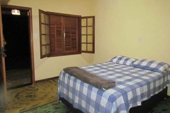 A imagem mostra um quarto simples da Pousada Fazenda Bocaina com uma cama de casal coberta com uma colcha de estampa xadrez em azul e branco. A parede é pintada em um tom amarelo claro. À direita da cama, há uma janela de madeira com venezianas abertas, permitindo a entrada de luz natural. Ao lado da janela, a porta do quarto também está aberta, revelando um pouco da área externa escura. No pé da cama, há uma toalha ou manta dobrada. O ambiente é básico, mas aconchegante, com um clima caseiro.