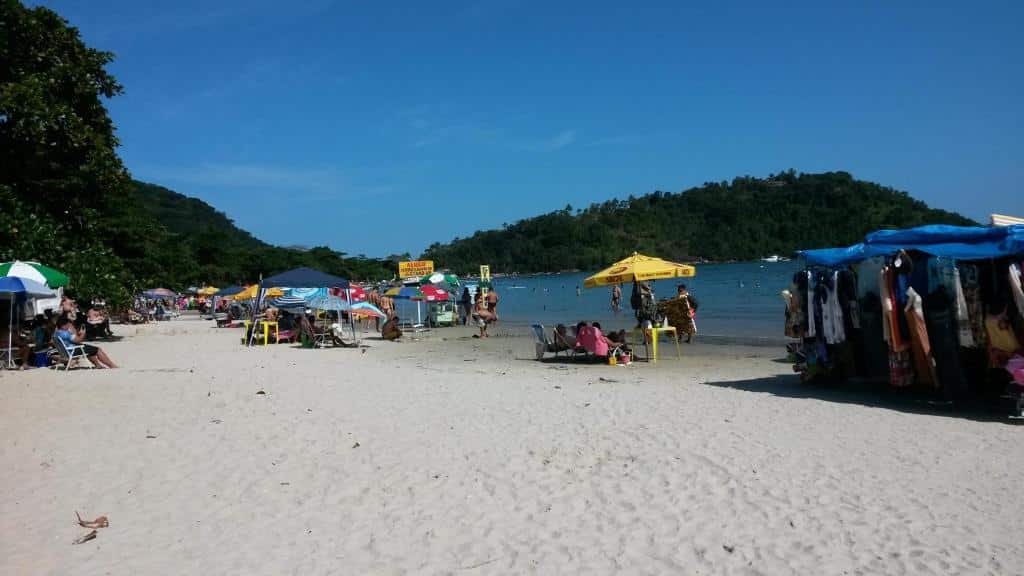 Praia do Lázaro em Ubatuba, com muitas pessoas na areia, guarda-sóis, o mar calmo e vegetação ao redor