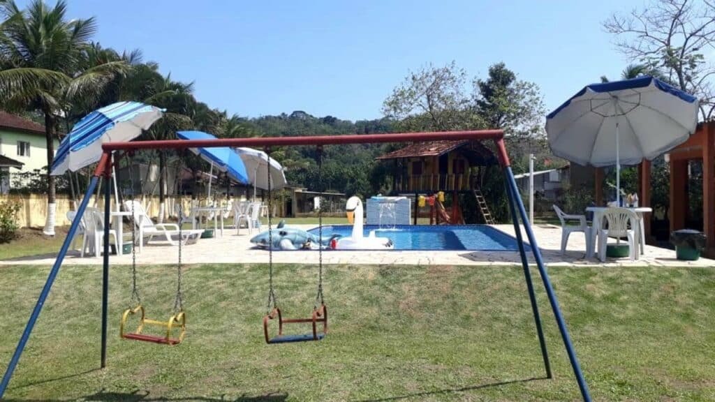 Área externa da Pousada Geres Peruíbe. Dois balanços na frente e atrás a piscina com boias e cadeiras de plástico ao redor. No fundo, um parquinho infantil de madeira.