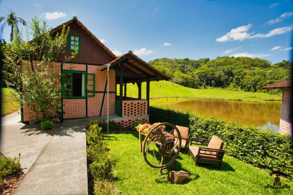 Parte da Pousada Imigrante que mostra um chalé com paredes de tijolos e um teto de madeira. Esse chalé tem uma varanda de frente para um lago e várias árvores em volta. Já do lado do chalé tem um gramado com duas cadeiras. Imagem ilustrando post Chalés em Pomerode.
