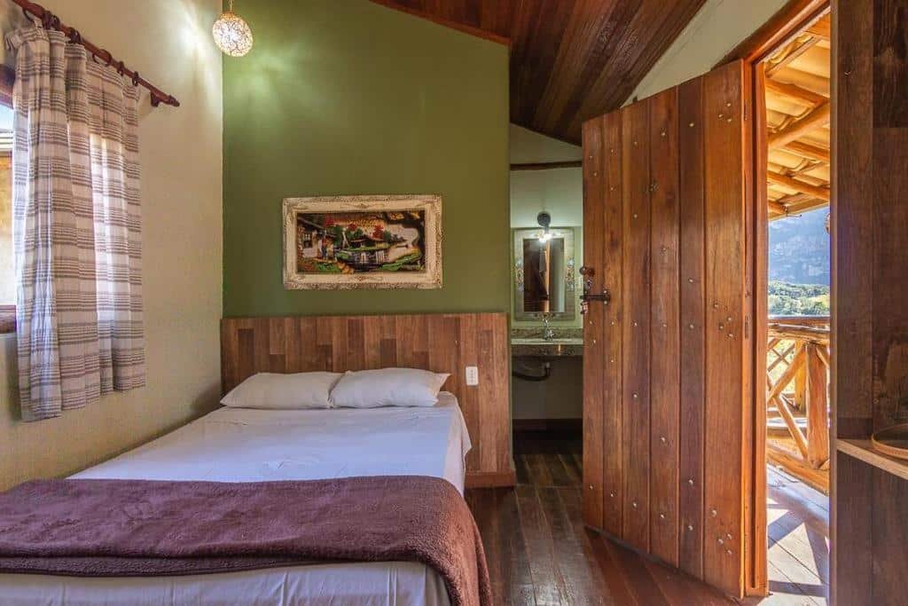 quarto da pousada lapralapa na Lapinha da Serra com uma cama de casal à esquerda, uma pia de banheiro com espelho ao fundo, no canto direito e uma porta de madeira maciça aberta na parede à direita mostrando a varanda e a natureza afora