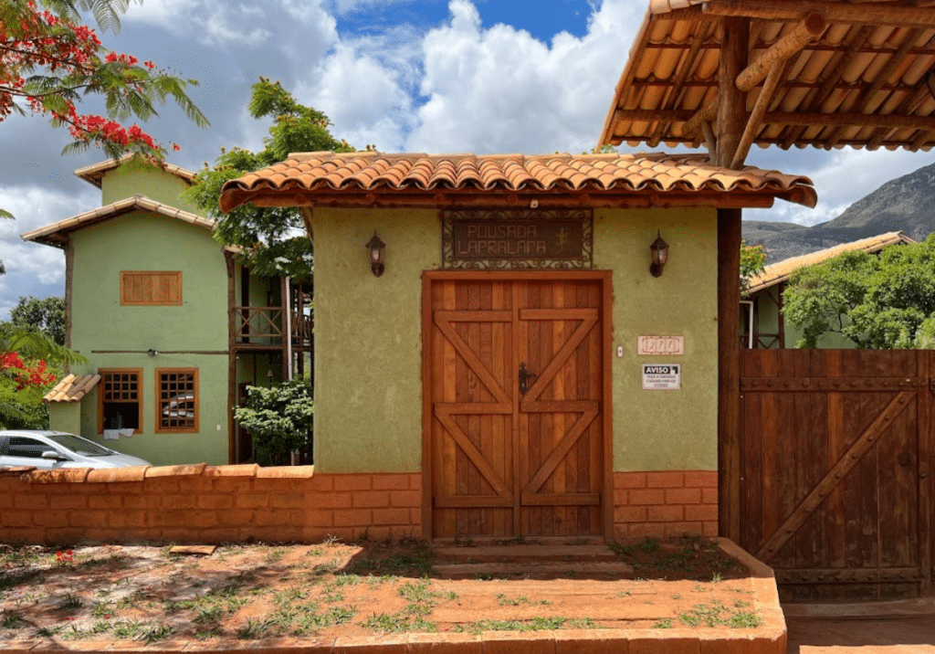 fachada da frente da pousada Lapralapa tirada pela Google Go Maps e postada online. A imagem apresenta a porta de entrada com o nome da pousada em cima da porta de madeira. Há duas lâmpada de cada lado da porta. Ao fundo é possível ver as montanhas e diversas árvores floridas.