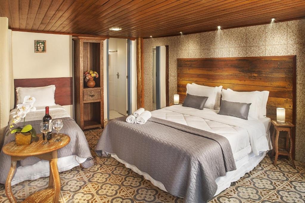 suíte da Pousada Makutta com uma cama de casal decorada com uma ampla cabeceira de madeira numa parede decorada com papel de parede estilizado. Há também uma pequena cama de solteiro e uma pequena mesa redonda com uma garrafa de vinho e duas taças de vidro.