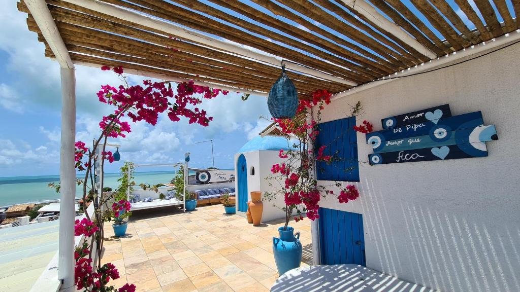 Área comum da Pousada Mediterrânea, em Pipa. Em primeiro plano está uma área com banco, janelas e flores, coberta com telhado de bambu. Ao fundo é possíver ver uma porta de entrada para o interior e, ao redor, sofás e espaços de descanso. Atrás de tudo isso está a praia.