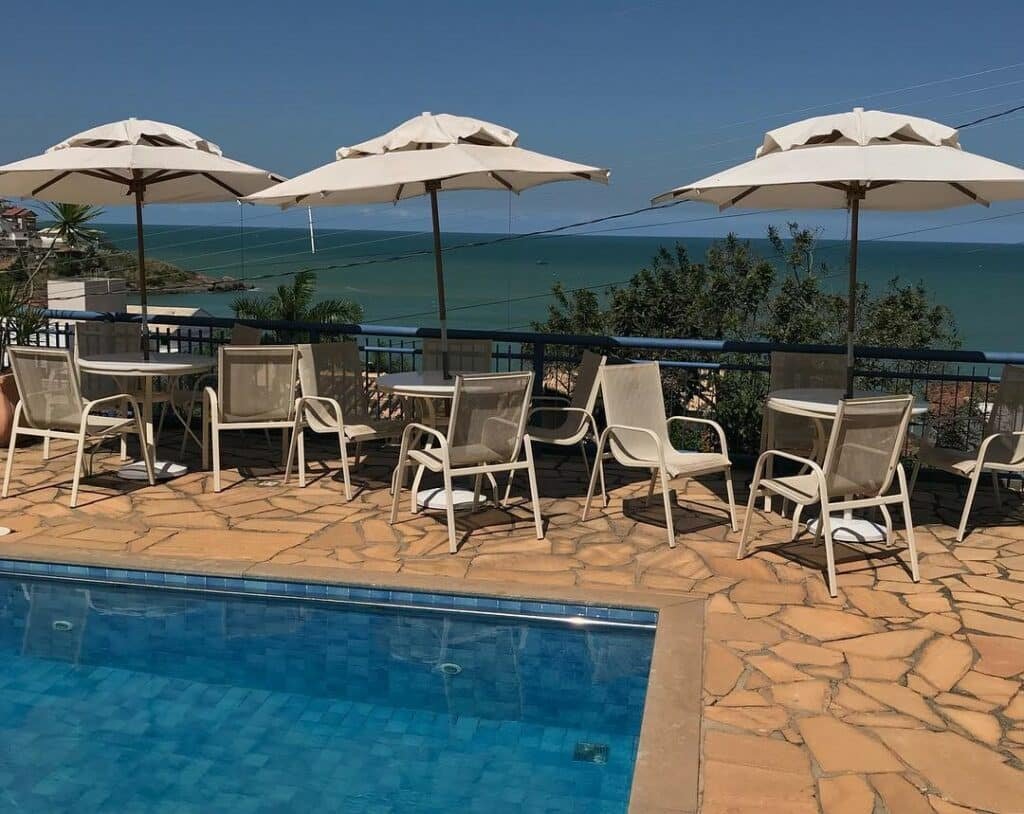 Piscina ao ar livre da Pousada da Memeia. Uma piscina na frente, atrás três mesas com cadeiras. Atrás uma grade de apoio e vista para o mar.