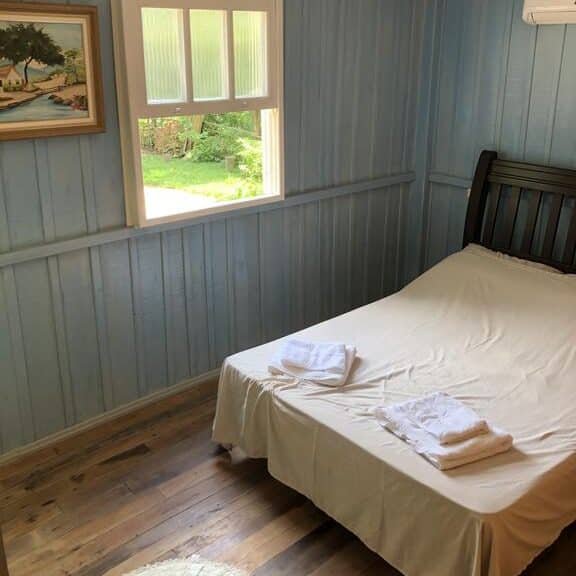 Quarto da Pousada Mestre Kikão, uma das pousadas na Serra do Rio do Rastro. Uma cama de casal está encostada na parede ao lado direito com toalhas dobradas aos pés. Uma janela na lateral do quarto tem vista para o jardim.