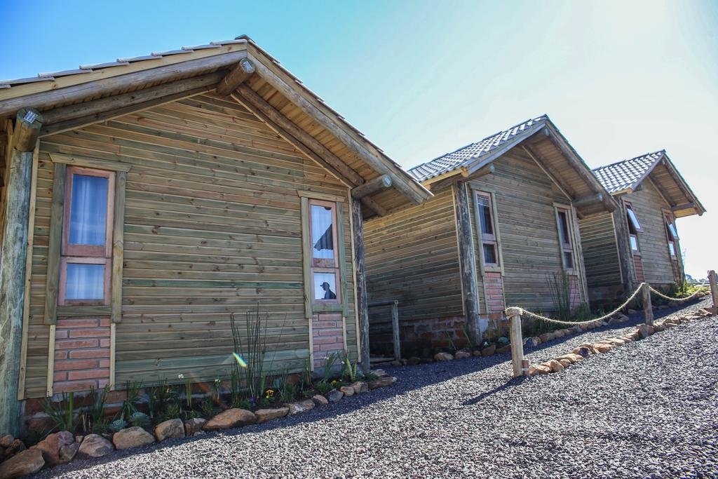 Três cabanas de madeira com detalhes em tijolos em um caminho de cascalho, sob um céu azul claro. Pequenas plantas margeiam o caminho.