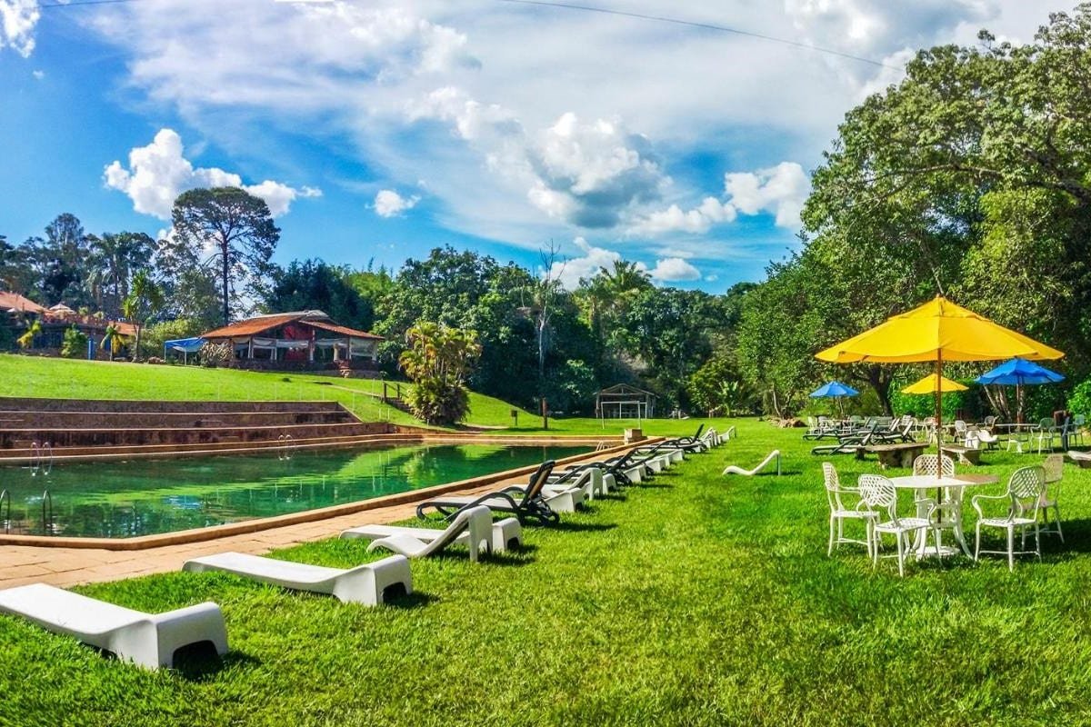 Área da piscina da Pousada Nascentes da Fortaleza, um dos hotéis fazenda em Minas Gerais, com espreguiçadeiras brancas, guarda-sóis amarelos e azuis e um gramado cercado por árvores. Céu azul claro acima.