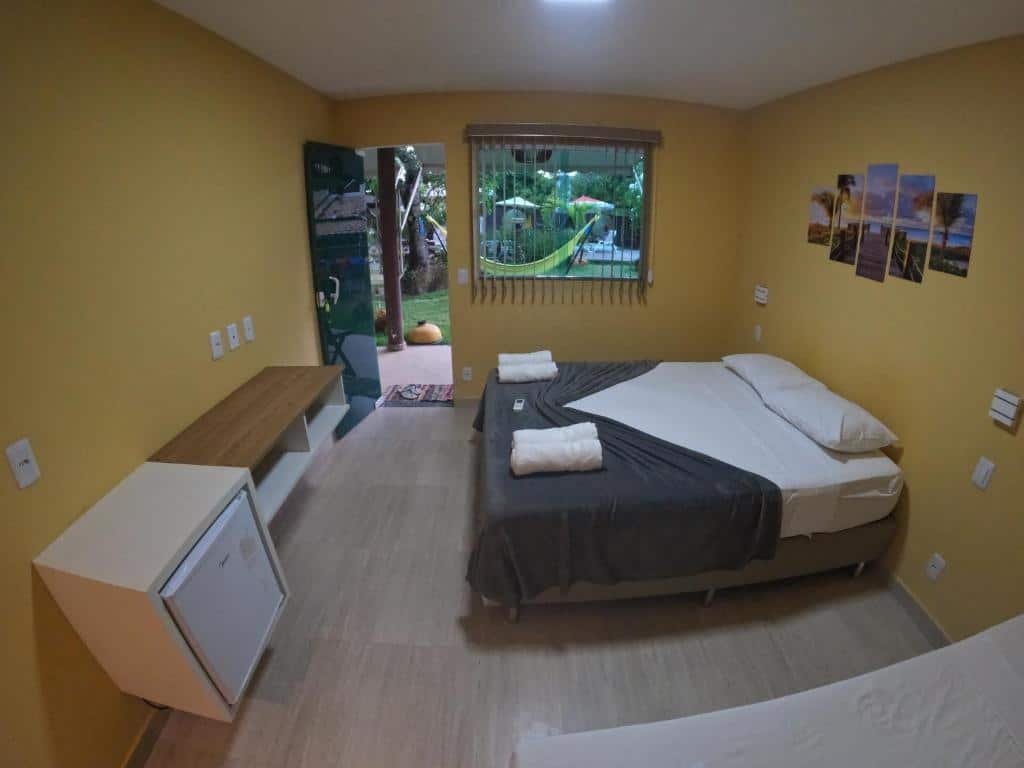 Quarto da Pousada Pedra da Lagoa Itacimirim. Uma cama de casal no lado direito, de frente um frigobar e uma prateleira, atrás a porta do quarto e a janela, com vista para o jardim.