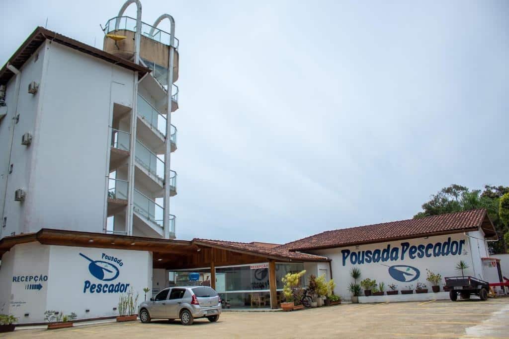 Entrada Pousada Pescador com um estacionamento aberto na frente, o hall de vidro e alguns vasos de flores perto da entrada
